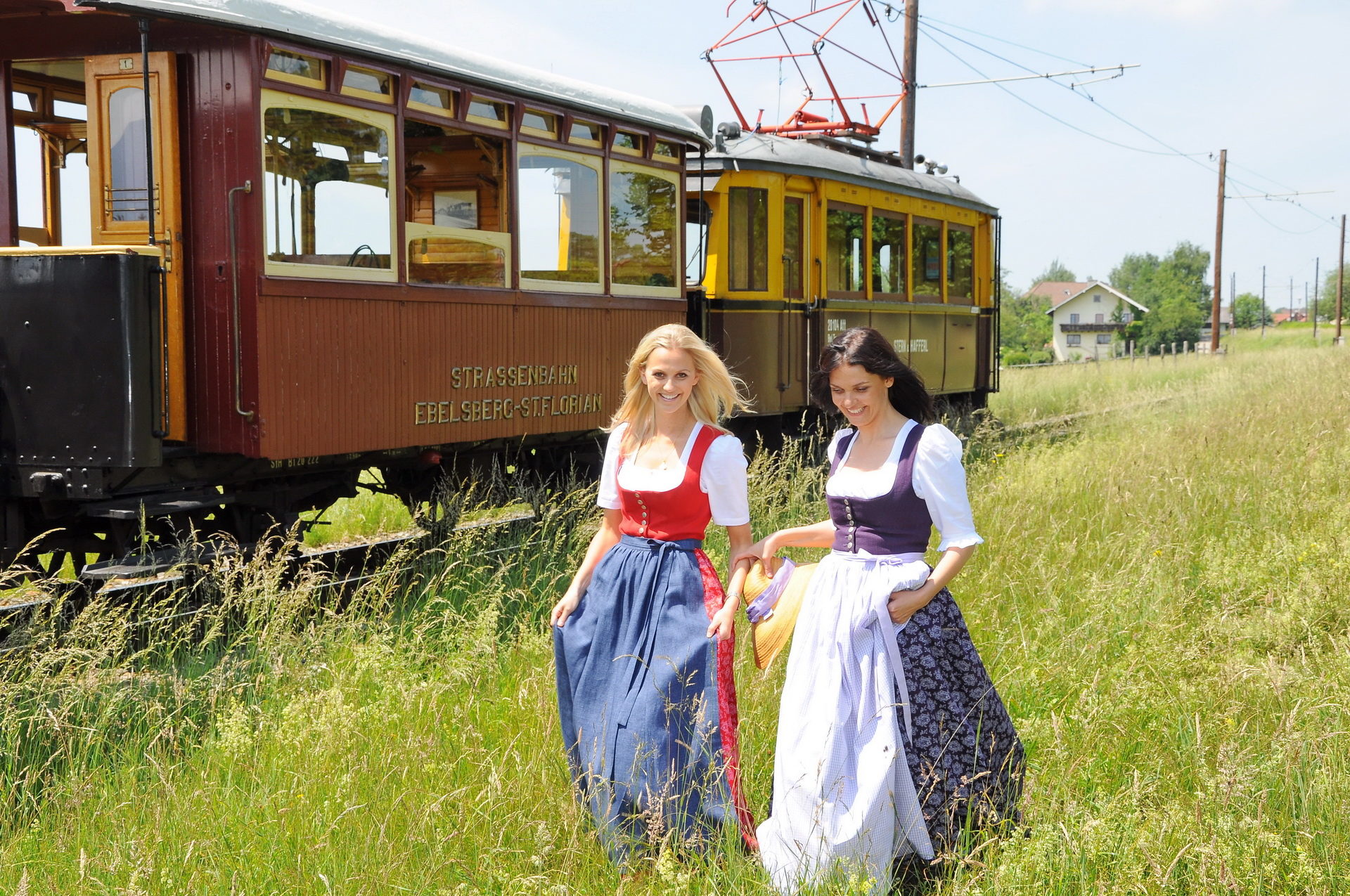Nostalgiefahrzeuge auf der Strecke der Atterseebahn mit 2 jungen Frauen im Vordergrund auf einer langen Wiese