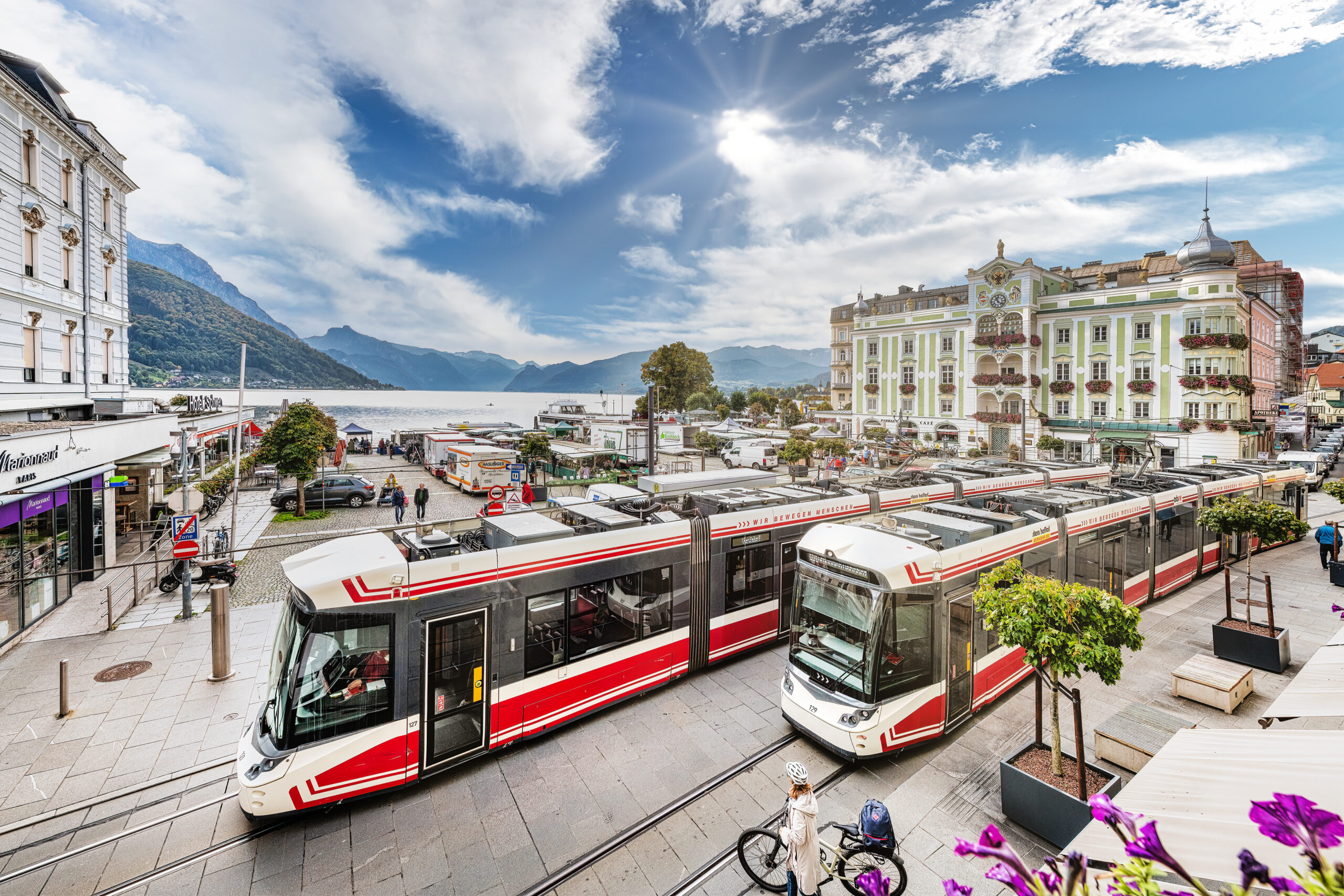 Foto vom Gmundner Rathausplatz mit 2 Tramlinks der Traunseetram im Vordergrund, Wochenmarkt am Rathausplatz und See und Berge im Hintergrund